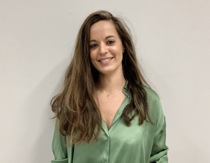 Eva. Pelo muy largo castaño claro con mechas rubias. Camisa escotada de seda verdes. Mira a casa sonriendo. De fondo, pared blanca.