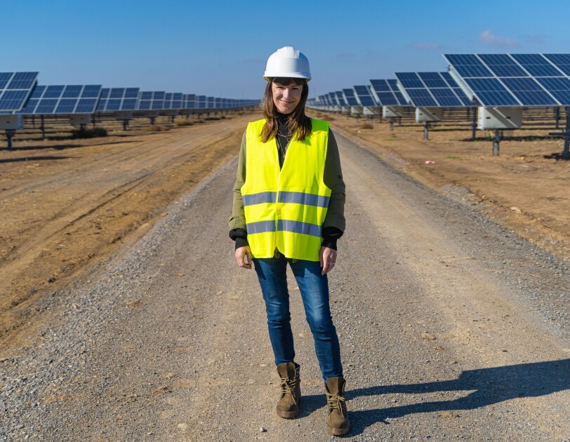 Denise Hugo, jefa de proyectos de Energías Renovables de Amazon. Ella está en medio de la foto, en un camino de tierra y entre dos líneas de placas solartes. Va vestida con unas botas marrones, unos vaqueros, casco blanco y chaleco amarillo.