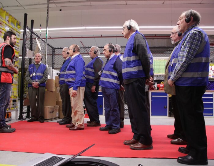 Nueve ingenieros jubilados durante la visita a un centro logístico. Todos ellos visten con un chaleco y escuchan atentamente las explicaciones del ingeniero que lleva un chaleco rojo.