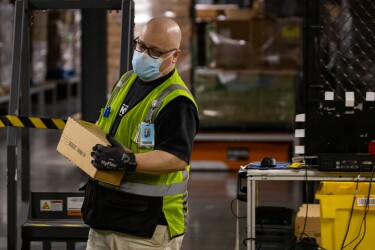A man wearing gloves, a mask, and a safety vest carries a small box.