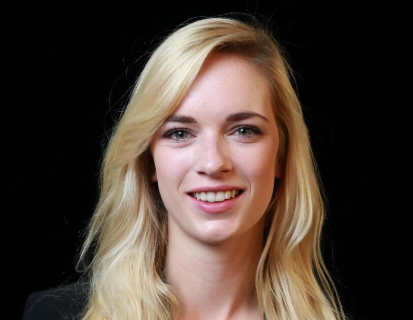 Eine Frau mit blonden langen Haaren und einem weißen T-shirt mit schwarzem Blaser schaut in die Kamera und lächelt.