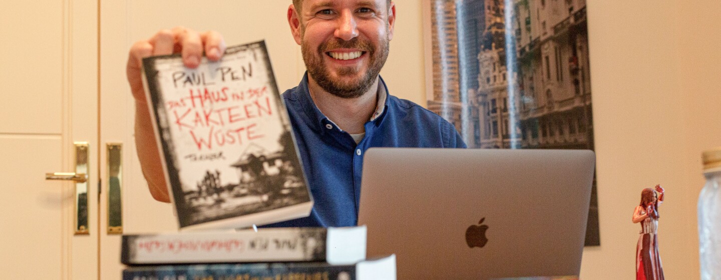 Paul Pen mostrando la traducción al alemán de su novela La casa entre los cactus. En su habitación de trabajo Paul sujeta su novela traducida al alemán con su mano derecha. Delante tiene el ordenador, sus otras novelas traducidas. De fondo hay una puerta y un poster colgado en la pared. Paul está mirando sonriendo a cámara con una camisa azul.