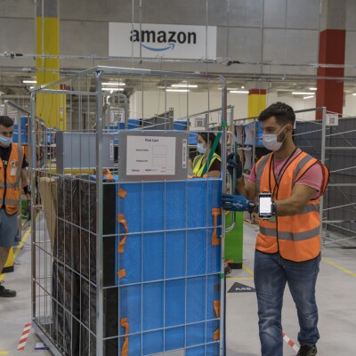 Un gruppo di dipendenti Amazon sposta dei carrelli contenenti dei pacchi. In alto sullo sfondo il logo di Amazon