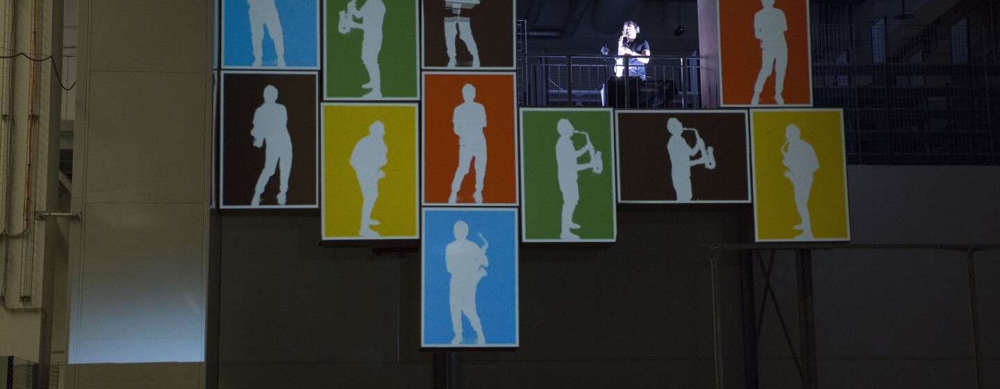 Video mapping realizzato al Centro di Distribuzione Amazon di Piacenza. Il disegno ricrea le sagome di un sassofonista in diverse posizioni, sullo sfondo di cubi colorati.