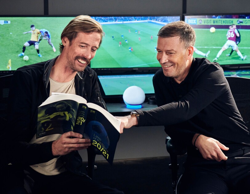 Peter Crouch (left) and Mark Clattenburg (right) reading through a football rule book in front of screens showing football games. On a table between them is the Amazon Echo Dot.
