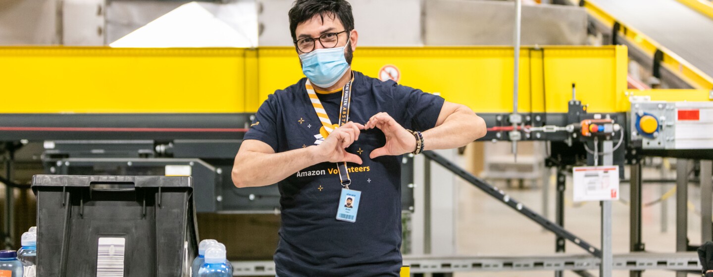 Ein Amazon Mitarbeiter mit Maske formt mit den Händen ein Herz. Auf dem T-Shirt liest man Amazon Volunteers.