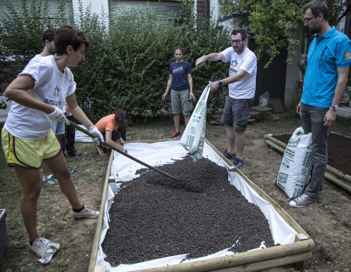 A scuola di verde: sei volontari Amazon all'opera nel giardino della scuola Media Leonardo da Vinci di Affori, nella periferia di Milano. Una donna in primo piano regge una pala con cui stende della terra, versata da un sacco da un uomo, in piedi di fronte alla donna. Gli altri 4 volontari osservano la scena.