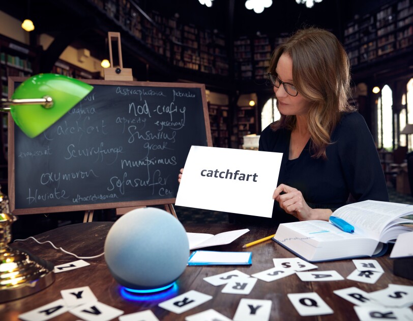 Susie Dent sitting at a library table next to a whiteboard holding a card that says the word "catchfart". On the table in front of Suzie there is an Amazon Echo and lots of letter cards.