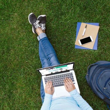 I woman types on the keyboard of a laptop while sitting in the grass.