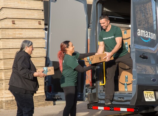 A man standing in an Amazon delivery van passes packages to two women standing on the ground. 