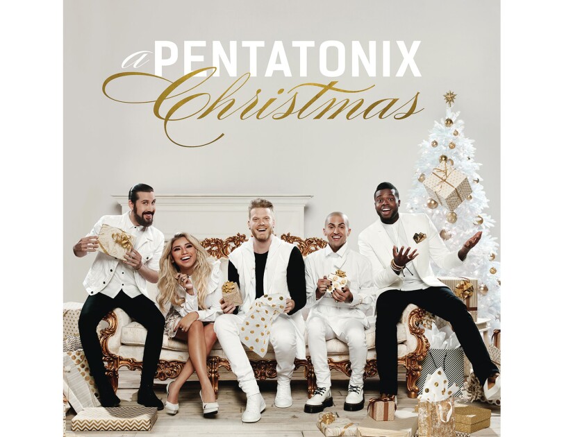 The members of Pentatonix sit on an oppulent ivory sofa with gold trim. A male in white button down with jacket and black pants sits next to a blonde woman leaning forward. She wears a white dress and flats. A man holds a gift, and wears white pants, black tee and white vest. A man in white tuxedo sits next to him and holds a gift. A man throws a gift up in the air. He wars a white tuxedo jacket with black and gold pocket square, white button down and tie and black pants.