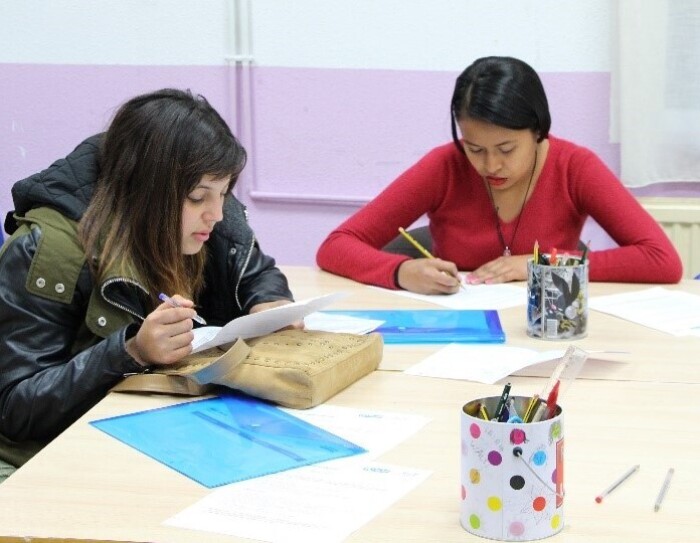 En una mesa blanca aparecen dos chicas estudiando. En la mesa hay hojas, dos carpetas azules y dos vasos con lápices y rotuladores. Una de las chicas lleva el abrigo puesto y lleva puesta una chaqueta negra y verde. Ella está sujetando una mano con un bolígrafo y en la otra tiene el papel. Está leyendo. El papel está sobre su bolso, que está encima de la mesa. Esta chica tiene el pelo oscuro y largo. La otra chica tiene un jersey rojo y está con un lápiz escribiendo en un papel.