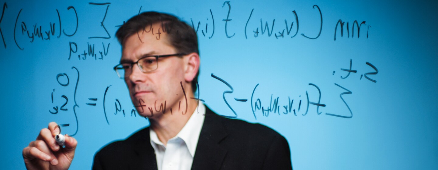 A man in a blazer and button-down shirt writes a complicated equation.