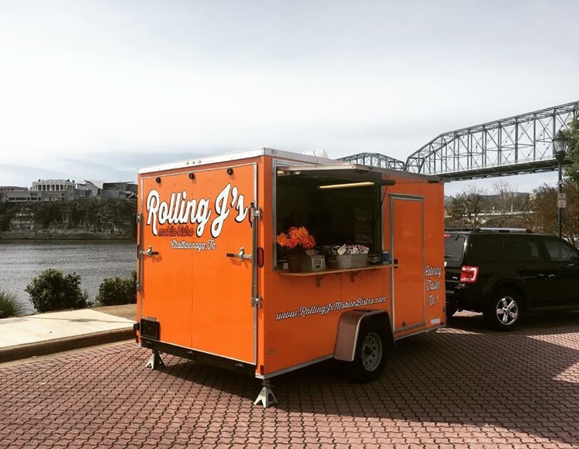 An orange food truck parked in front of the Tennessee River. The food truck has "Rolling J's" branding on it, and is parked on a street made of pavers.