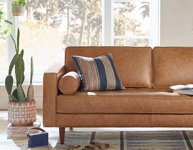 A geometric planter holds a large cactus, and sits atop several coffee table books. Next to it is a leather midcentury sofa, topped with a striped pillow. The sofa and planter both sit on a faded geometric rug.