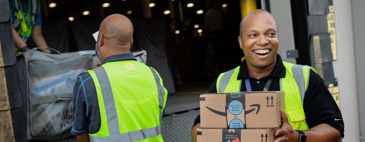 A smiling man holds boxes while people in the background load a truck. 