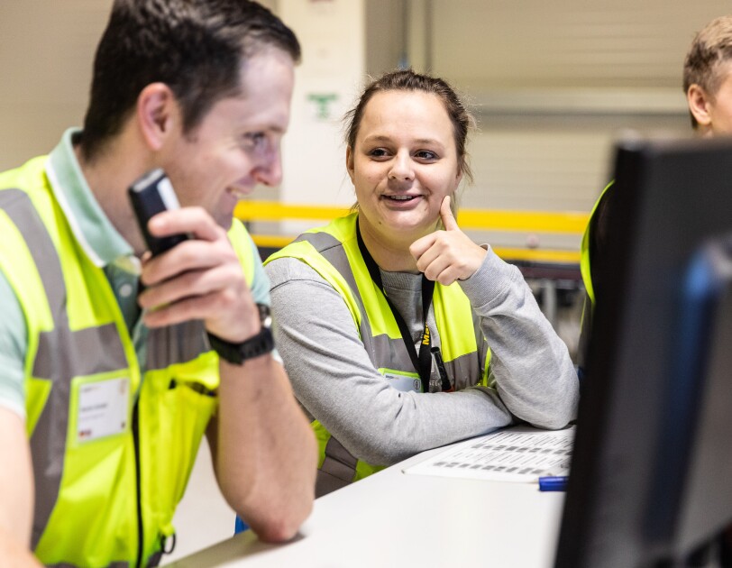 Amazon Logistik Mitarbeiter im FC, sie tragen gelbe Sicherheitswesten, sie stehen am Schreibtisch vor Bildschirmene und unterhalten sich