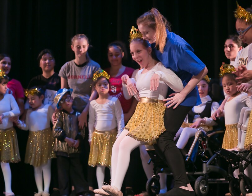 A dancer wearing ballet slippers, a gold skirt, a tiara, and a white leotard and tights performs. The dancer has cerebral palsy and is leaning back against a helper for support. Other dancers and helpers are in the background.
