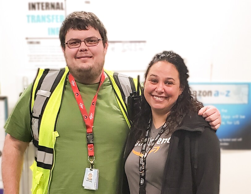 A woman and a man pose together for a photograph. The man wears a green T-shirt and a yellow safety vest. The woman wears a black fleece jacket and a gray T-shirt with the Amazon smile logo.