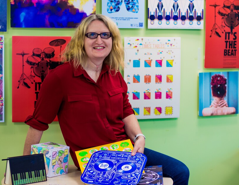 A blonde woman wearing glasses, jeans, and red button up shirt is seated on a tabletop in a room painted green, with myriad artwork hung on the wall.