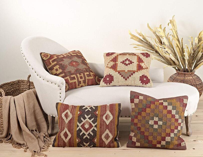 Four kilim-patterned pillows in earth tones rest on and around a chaise lounge. A basket with a cocoa fringe blanket sits to the left of the chaise, and a woven planter with decorativ dried branches sits to the right.