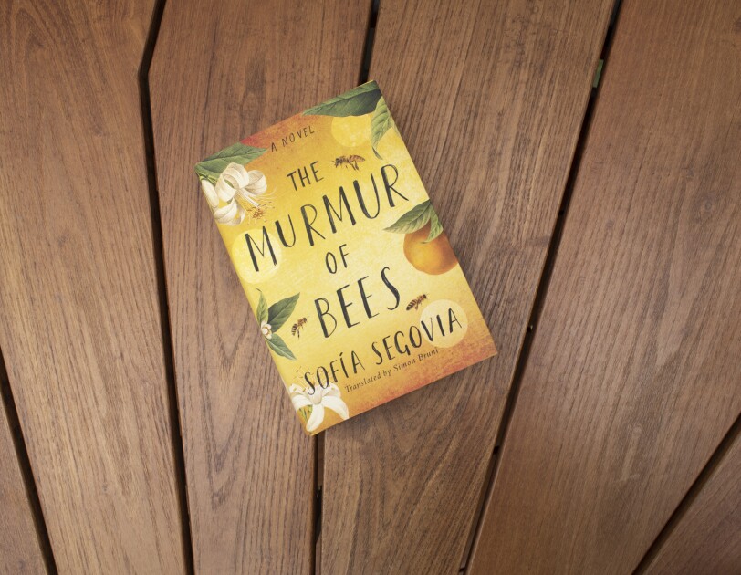 Book cover for "The Murmur of Bees" on a teak wood background.