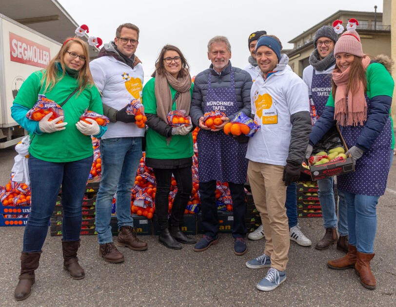 Eine Gruppe von Amazon Mitarbeitern, u. a. von Fresh mit grünen Shirts, der Rest mit weißen Shirts, steht vor einem Berg von Orangen und Mandarinnen. In ihrer Mitte: Paul Breitner.