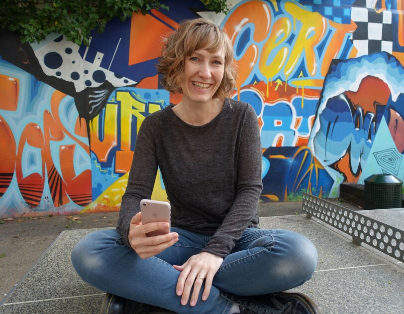 Eine Frau mit kurzen blonden Haaren und einem grauen Pullover sitzt auf einer Tischtennisplatte und lacht in die Kamera.
