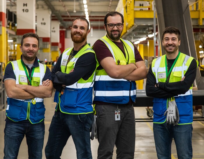 Quattro uomini con gilet ad alta visibilità e le braccia incrociate, guardano in camera e sorridono. Sono nel centro di distribuzione Amazon di Passo Corese.