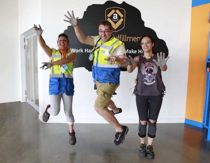 Patricia Alonso, Maribel Salcedo y Marius Dumitrescu aparecen en la entrada del centro logístico saltando y mostrando las palmas de sus manos pintadas de color gris. Están sonriendo mirando a cámara. Marius se enceuntra en medio.