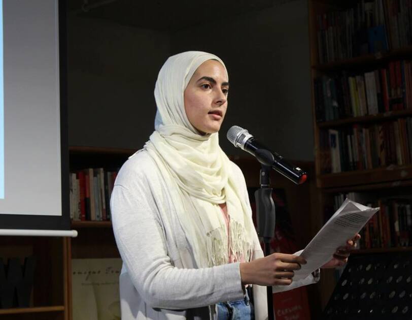 A woman (Sumaya Awad) stands in front of a microphone, speaking about her experience