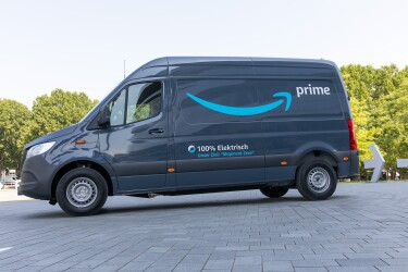 Una furgoneta eléctrica de Mercedes-Benz con el logo Prime de amazon y debajo en alemán 100% eléctrica. La furgoneta sin conductor está en una plaza con árboles en el fondo.
