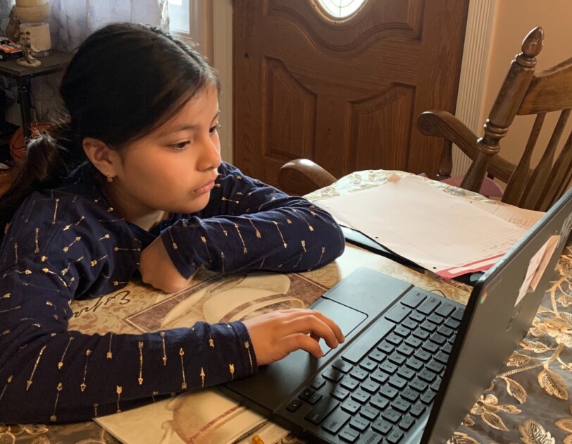 A girl sits a dining room table with a tablecloth and works on a laptop computer.