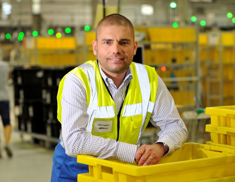 Ein Mann in gelber Sicherheitsweste lehnt sich lässig auf einem Stapler gelber Transportbehälter im Logistikzentrum