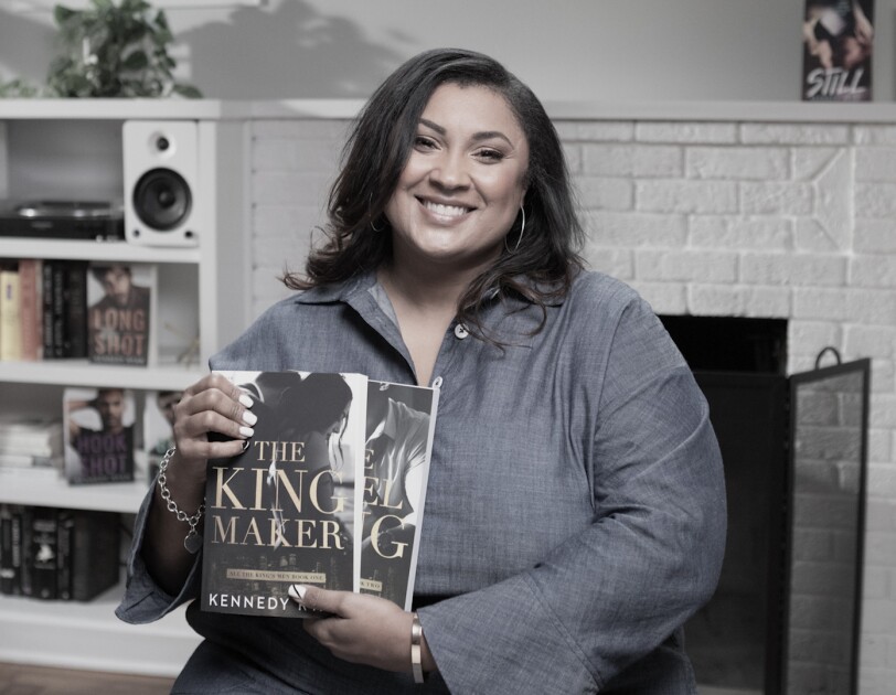 A woman sits in a living room setting, holding two books she wrote and self-published.