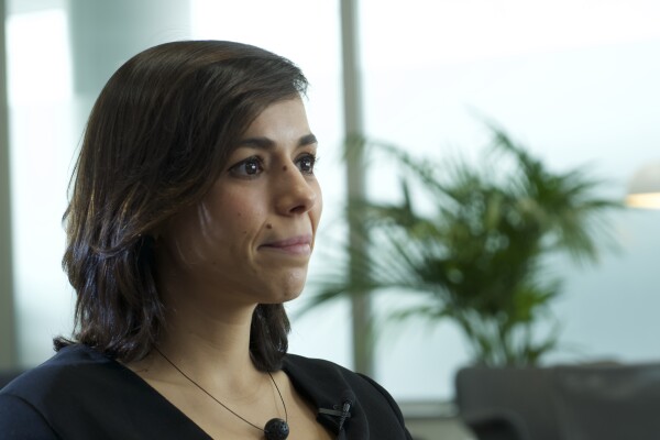 Natalia Gutiérrez, Líder Global de Impacto Sostenible y Seguridad de Cabify. De fondo unas butacas de color gris con plantas verdes y una lámpara de pie de color blanco. Unos ventanales grises grandes. La protagonista en primer plano con una camisa cruzada negra de cuello pico, manga larga y botones de color blanco. Con el pelo castaño, liso hasta el hombro y sonriendo mirando a la derecha de la foto. Lleva un collar de alambre con una bola. 