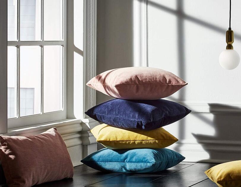 A stack of four pillows sit on a wood floor. Pillows are (from bottom) peacock blue, ocre, indigo and dusty pink. They sit in front of a low, paneled window, and besides another pink pillow on the left, and an ocre one on the right.
