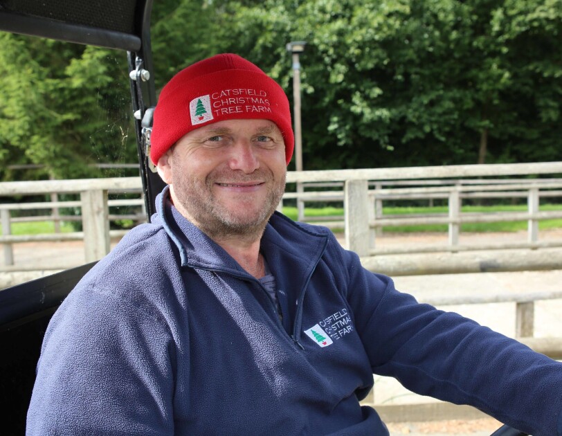 Clive Collin, owner of Catsfield Christmas Tree Farm, sitting in a buggy outside of his Christmas tree farm. He is wearing a beanie hat with his company logo on it.