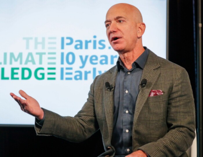 De fondo el cartel the The Climate Pledge, Paris 10 years early. Jeff Bezos en primer plano con una mano levantada. Una amerciana verde y gris a cuadros y una camisa azul. En la americana lleva un pañuelo rojo.