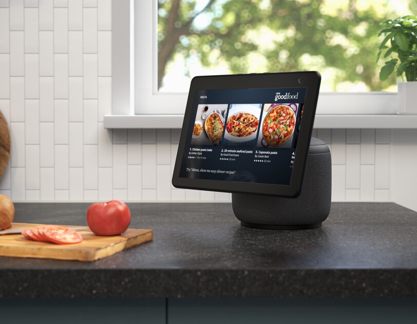 An image of the new Echo Show 10 on a kitchen counter. The device is showing recipes on BBC goodfood and next to the device there is a chopping board with tomatoes and onions.