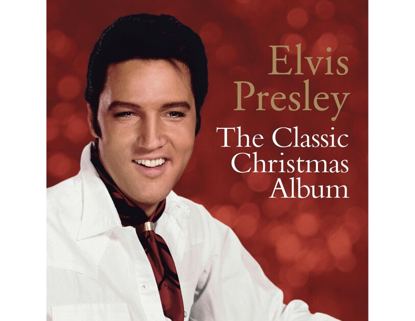 Elvis Presley wears a white button down and burgundy neck kerchief with a gold clasp. Behind him, we see out-of-focus lighting against a red background.
