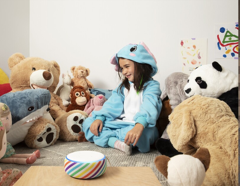 A child sits in a playroom, surrounded by stuffed animals. In front of her is an Echo Dot device is a rainbow stripe, on a tabletop.