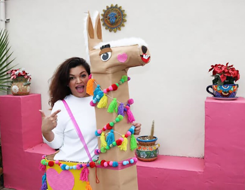 A woman in her "llama boxtume" - a costume made of Amazon boxes, with a larger box as the llama body, a tall box for the neck, and a smaller box for the head, the llama has felt details for eyes, nose and mouse, and is wrapped in decorations made of pom poms and tassels.