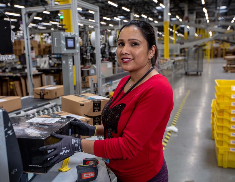 An Amazon associate holds a product near a conveyor belt. Tools sit on the countertop near her. She wears a red long-sleeve tee shirt and work gloves as she works at a Columbus, Ohio fulfillment center, CMH1
