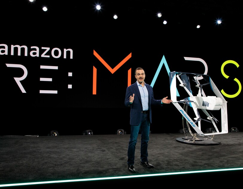 Jeff Wilke, CEO consumer worldwide at Amazon, stands on a stage. Behind him is a re:MARS logo. To his left is a drone painted in white and light blue.