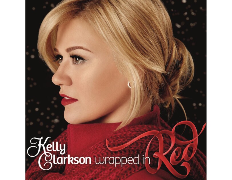 Kelly Clarkson wears her blonde hair tied behind her head, a red high-collared shirt with red scarf around her shoulders. Silver earrings, eye liner, and red lipstick completes her look. She looks to her right.