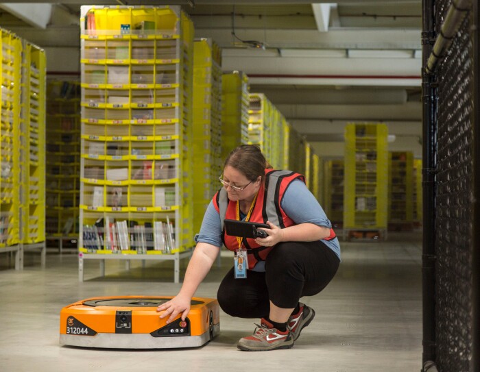 Foto di Maria Stella Carnà che regge un tablet di controllo e si china su uno dei robot di cui è responsabile all'interno del Centro di Distribuzione Amazon di Passo Corese