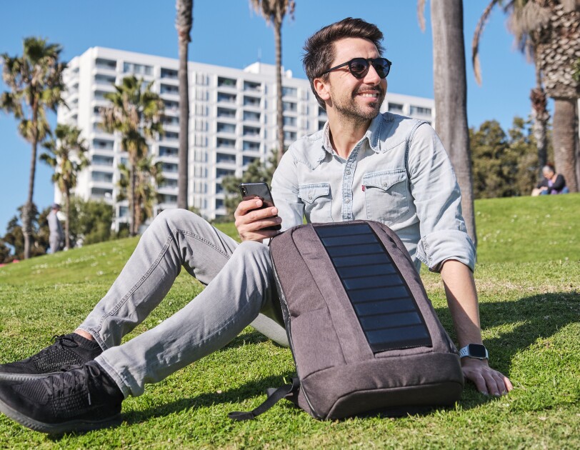 Ein Mann mit braunen Haaren und einem blauen Hemd sitzt auf einer Wiese. Neben sich hat er einen grauen Rucksack mit Solarpannels.