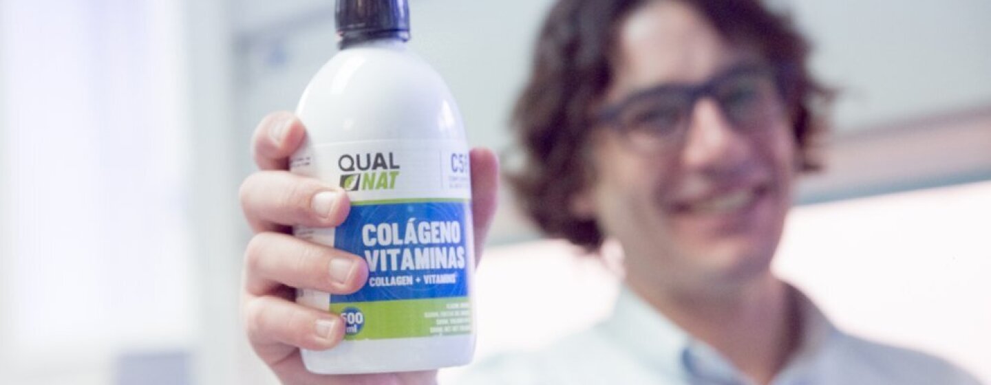 Carmelo Castro de Qualnat mostrando el bote de colágeno. Él, que se encuentra en el laboratorio, esta desenfocado de fondo con una camisa blanca.