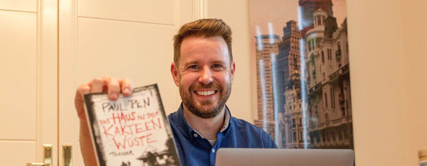 Paul Pen mostrando la traducción al alemán de su novela La casa entre los cactus. En su habitación de trabajo Paul sujeta su novela traducida al alemán con su mano derecha. Delante tiene el ordenador, sus otras novelas traducidas. De fondo hay una puerta y un poster colgado en la pared. Paul está mirando sonriendo a cámara con una camisa azul.